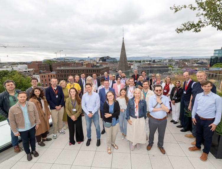 Large delegation gathered on RCSI rooftop terrace overlooking a city skyline and church spire under a cloudy sky.