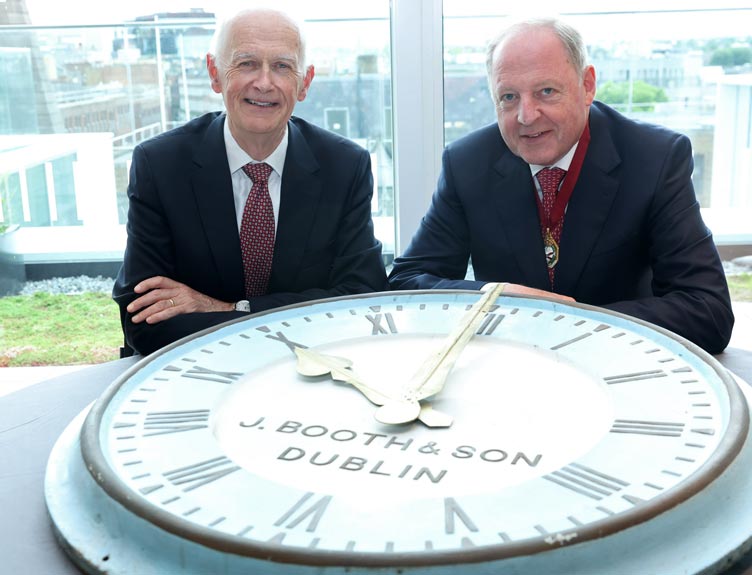 RCSI Vice Chancellor and President in dark business suits sit behind a large historic clock face displayed on a table.