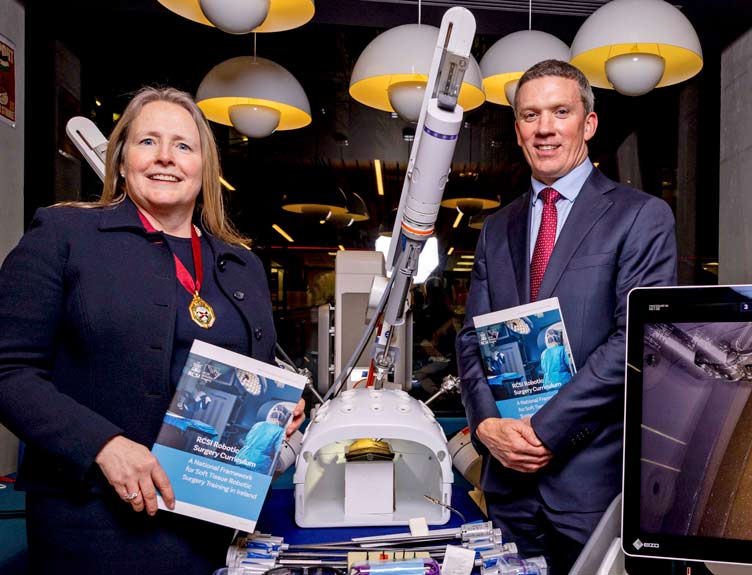 Two people hold printed copies of RCSI's National Robotic Surgery Curriculum surrounded by robotics