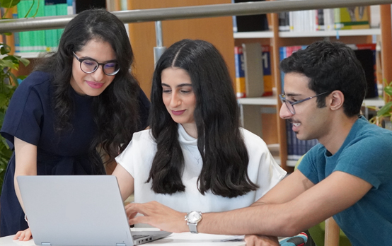 Three students collaborating around a laptop in a library, reviewing information together at a study table with bookshelves in the background.