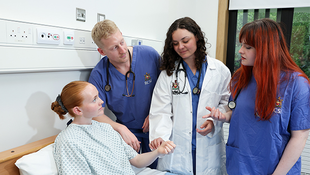 A tutor and group of RCSI med students in white coats and scrubs take a patient's pulse
