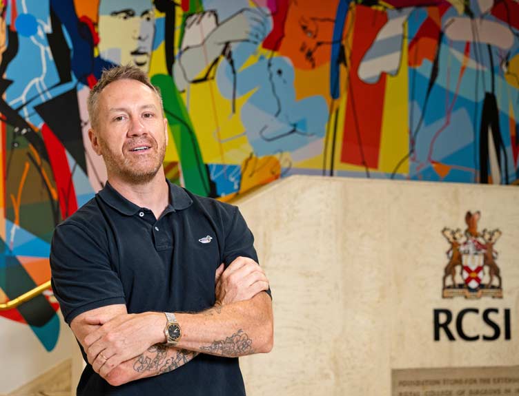 Artist stands in front of large scale colourful artwork on university wall