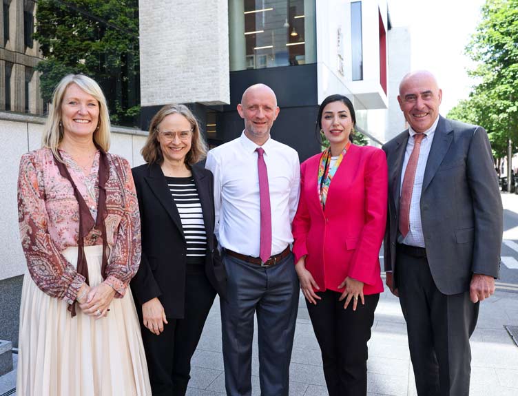Five adults in business attire stand side-by-side outdoors in front of a modern building, facing the camera.