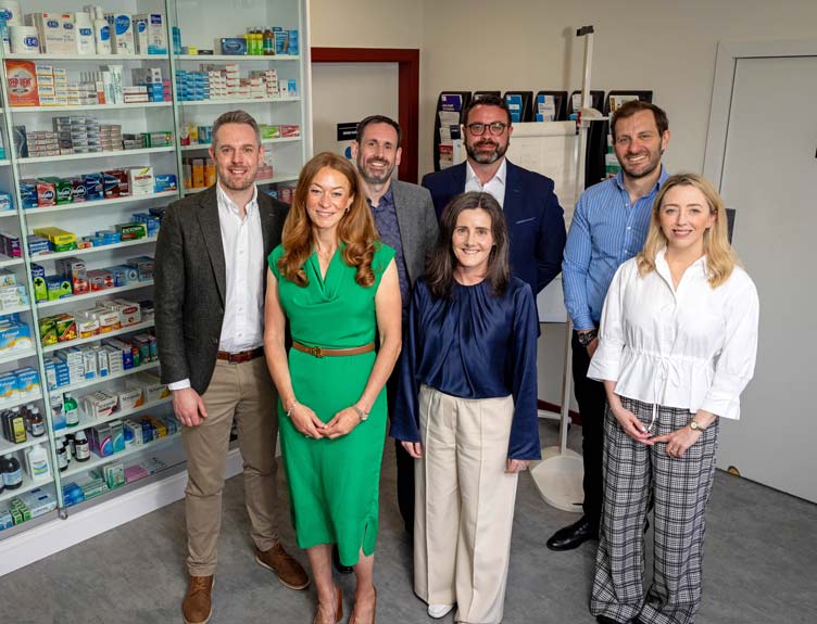 Seven RCSI and Uniphar team members stand together inside a pharmacy or medical retail setting, posed in front of shelves stocked with medicines and health products.