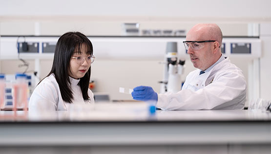 Academic staff member demonstrating a laboratory technique to a student or junior staff member in a biomedical research laboratory, with both wearing laboratory coats and safety glasses.