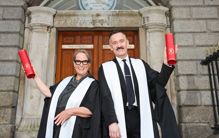 Dr Diana Mason and Mr Brendan Courtney in graduation gowns stand outside a historic stone building with large wooden doors, each holding a red cylindrical degree scroll case.