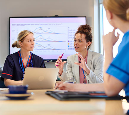 Three managers and healthcare professionals at a table around a laptop, with a big-screen presentation in the background.